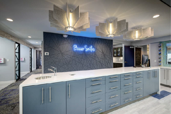 A modern dentist office reception area with a blue-themed desk, neon sign saying "bauer smiles", geometric wall pattern, hanging geometric lights, and a clean, stylish interior design.