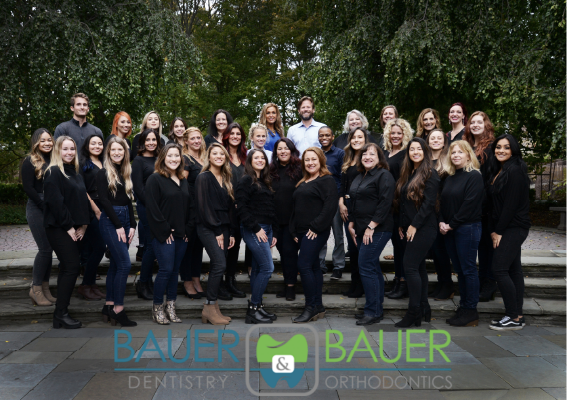 A group of dentist and orthodontics staff, primarily women, posing outdoors in a lush garden setting, dressed in black. The Bauer Dentistry & Orthodontics logo is visible at the bottom.